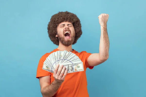 Portrait of extremely happy satisfied man with Afro hairstyle wearing orange T-shirt holding dollar bills and clenched fist, received an inheritance. Indoor studio shot isolated on blue background.