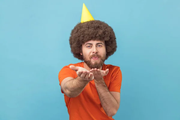 Portrait of romantic man with Afro hairstyle in orange T-shirt and party cone sending air kisses to his guests, looking at camera, celebrating birthday. Indoor studio shot isolated on blue background.