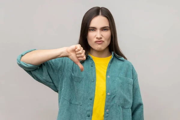 Dissatisfied woman showing thumb down, disapproval sign, gesturing dislike to bad service, disagree with suggestion, wearing casual style jacket. Indoor studio shot isolated on gray background.