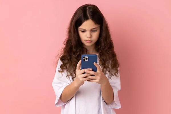 携帯電話上のソーシャルメディアで白いTシャツのテキストメッセージを着て深刻な少女の肖像画 モバイルネットワークサービスを使用して オンラインでチャット ピンクの背景に隔離された屋内スタジオショット — ストック写真