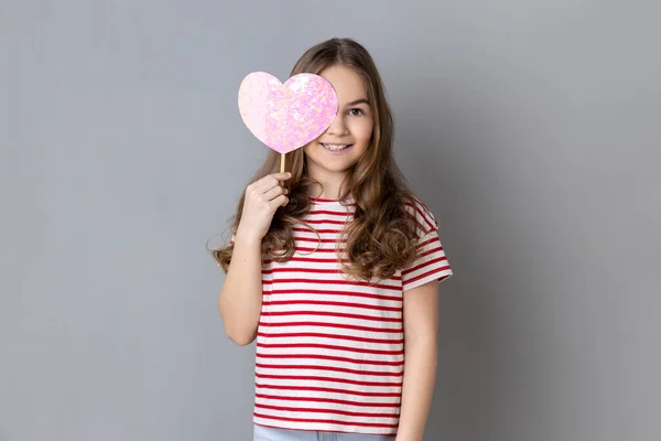 満足した笑顔の少女の縞模様のTシャツを着ての肖像画の棒にピンクの心で彼女の目をカバーし ロマンチックな感情を示しています 灰色の背景に隔離された屋内スタジオショット — ストック写真