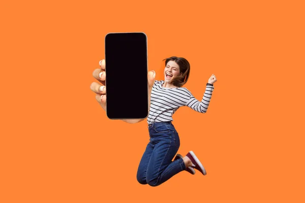 一个极其快乐快乐的女人的画像 她紧握拳头跳起来 庆祝她的成功 用空屏幕显示手机 在橙色背景下被隔离的室内拍摄 — 图库照片