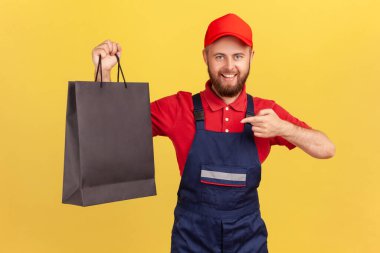 Mavi üniformalı mutlu kurye portresi siyah alışveriş çantasını gösteriyor müşteriye sipariş veriyor dişlek gülümsemeyle kameraya bakıyor. Sarı arka planda kapalı stüdyo çekimleri