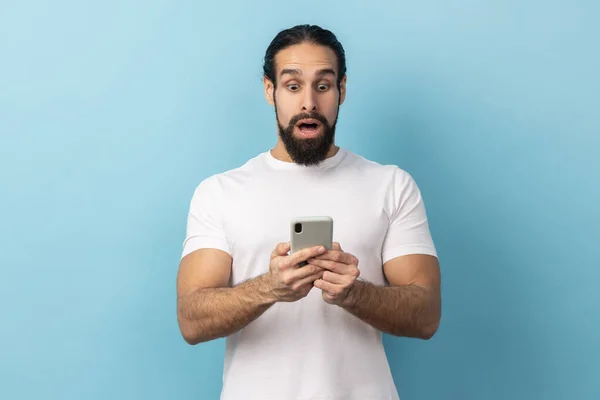 身穿白色T恤的大胡子男子的画像 一边用手机 一边上网 一边是惊人的消息 室内拍摄被蓝色背景隔离 — 图库照片