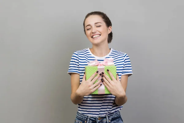 快乐微笑的女性形象 身穿条纹T恤 抱着绿色包装的礼品盒 心满意足 在灰色背景下被隔离的室内拍摄 — 图库照片
