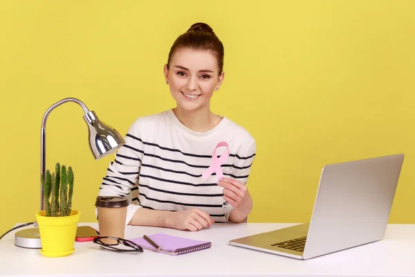 職場でノートパソコンを持って座ってピンクのリボン 乳がんの認知度 女性の健康の象徴を示す楽観的なきれいな女性のボランティア 黄色の背景に隔離された屋内スタジオショット — ストック写真