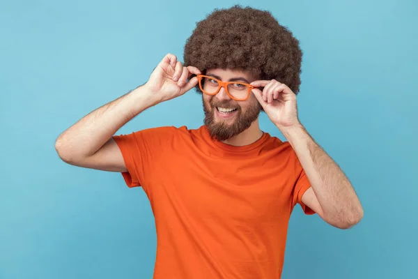 Delighted satisfied man with Afro hairstyle in orange T-shirt admires eyeglasses, standing in a new spectacles, looking at camera with toothy smile. Indoor studio shot isolated on blue background.