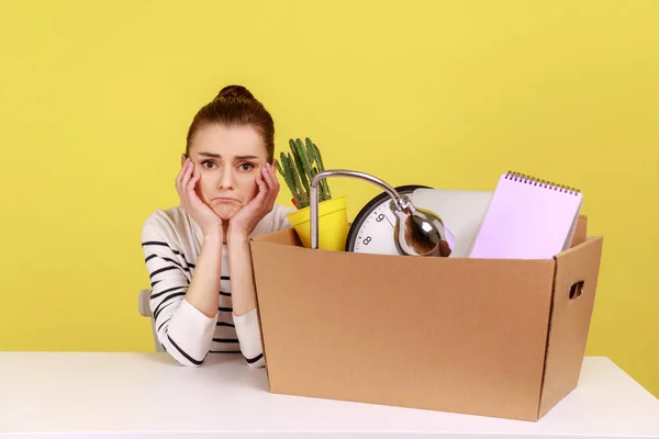Unhappy depressed woman office worker sitting at workplace with cardboard box with her things, dismissal, bankruptcy, keeps hands under chin. Indoor studio studio shot isolated on yellow background.