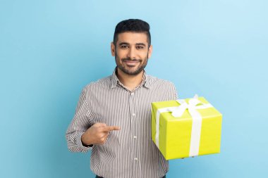 Sakallı mutlu iş adamı, elinde sarı hediye kutusu, parmağıyla kameraya bakıyor, dişlek gülümsemeli, çizgili gömlek giyiyor. Mavi arka planda kapalı stüdyo çekimi.