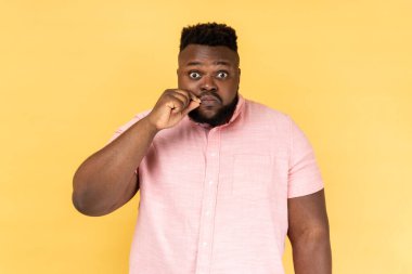 Kimseye anlatmam. Gözü korkan, pembe tişörtlü, fermuarlı dudaklı, kameraya bakan, ağzı kapatan ve sır saklayacağına söz veren bir adamın portresi. Sarı arka planda kapalı stüdyo çekimleri.