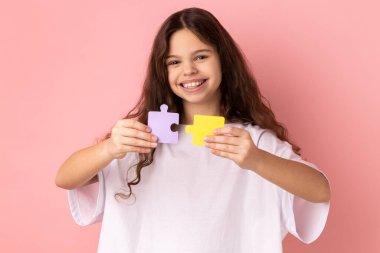 Beyaz tişört giyen, elinde sarı ve mor yapboz parçaları tutan mutlu küçük kız portresi çözme görevi, kameraya bakma. Pembe arka planda kapalı stüdyo çekimleri.