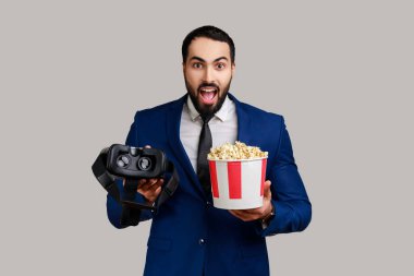 Şaşkın sakallı adamın elinde sanal gerçeklik gözlüklerini kullanarak sanal gerçeklikte patlamış mısırlı film izlemesine şaşırdım. Gri arka planda izole edilmiş kapalı stüdyo görüntüleri.