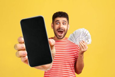 Kırmızı tişörtlü, sakallı, ağzı açık, ağzı açık, ağzı açık, elinde para tutan ve cep telefonunu boş ekranla gösteren mutlu bir adam. Sarı arka planda kapalı stüdyo çekimleri.