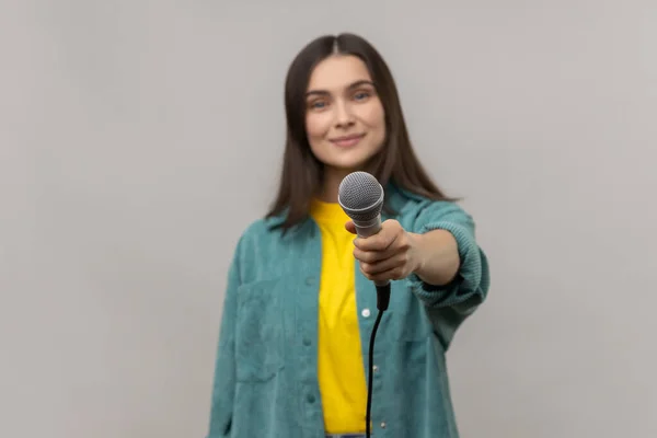 Portrait of satisfied woman with dark hair standing offering microphone, journalist or reporter taking interview, wearing casual style jacket. Indoor studio shot isolated on gray background.