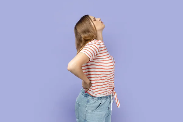 Side view of blond woman wearing striped T-shirt touching back and shouting from pain, suffering back ache, having discomfort lower lumbar muscular. Indoor studio shot isolated on purple background.