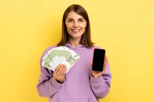 肯定的な満足暗い髪の女性が空の画面でユーロ紙幣や携帯電話を保持し オンライン賭け 紫色のパーカーを着て 黄色の背景に隔離された屋内スタジオショット — ストック写真