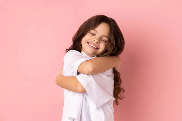 満足した幸せな女の子の白いTシャツを着て自分自身を受け入れるの肖像画 目の喜びと幸福から閉じ立っている ピンクの背景に隔離された屋内スタジオショット — ストック写真