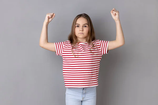 ストライプのTシャツを着た強い暗い髪の少女が腕を上げて立ち上がり 彼女の強さを示し 真剣な表情でカメラを見ています 灰色の背景に隔離された屋内スタジオショット — ストック写真