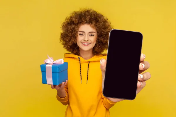 Cute delighted woman with Afro hairstyle wearing casual style hoodie holding present box and showing big empty cell phone display for advertisement. Indoor studio shot isolated on yellow background.