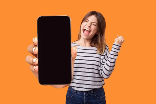 快乐快乐的女人穿着条纹衬衫尖叫的画像 快乐地展示手机广告模型区 庆祝她的胜利 在橙色背景下被隔离的室内拍摄 — 图库照片