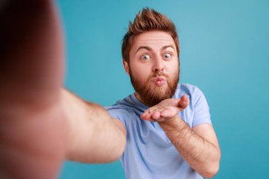 Romantik flört portresi sakallı yakışıklı blog yazarı özçekim POV 'u yapıyor, takipçilerine canlı yayın yaparken hava öpücüğü gönderiyor. Mavi arka planda kapalı stüdyo çekimi.