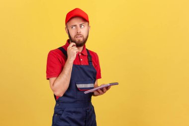 Mavi tulumlu, elinde kağıt defteriyle duran, emirleri yazan düşünceli bir yüz ifadesi olan dalgın işçi portresi. Sarı arka planda kapalı stüdyo çekimleri.