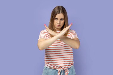 Üzerinde çizgili tişört olan sıkı sarışın bir kadının portresi X işareti taşıyor, jest yok, kameraya patronluk taslayan bir ifadeyle bakıyor. Mor arka planda izole edilmiş kapalı stüdyo çekimi.