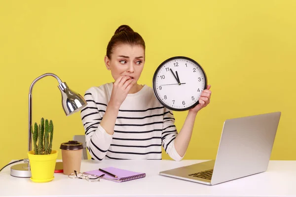 Nervous unhappy woman office worker biting finger nails, holding wall clock talking video call on laptop sitting at workplace, time to break. Indoor studio studio shot isolated on yellow background.