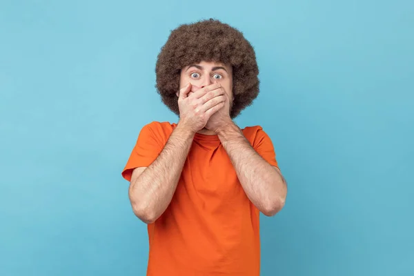 Man with Afro hairstyle in orange T-shirt closing mouth not to scream, feeling frightened and terrified, his eyes and look full of fear and terror. Indoor studio shot isolated on blue background.