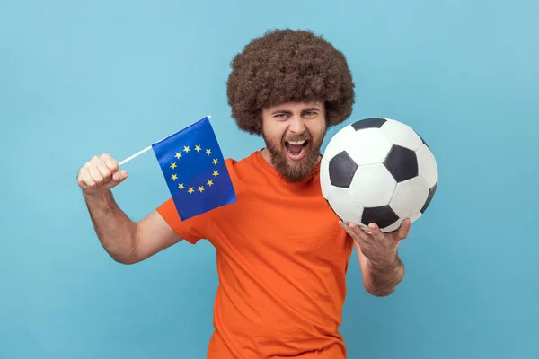 穿着T恤衫 头戴足球和欧盟旗帜的非洲式发型的人非常快乐 为他最喜欢的球队欢呼 庆祝胜利 室内拍摄被蓝色背景隔离 — 图库照片