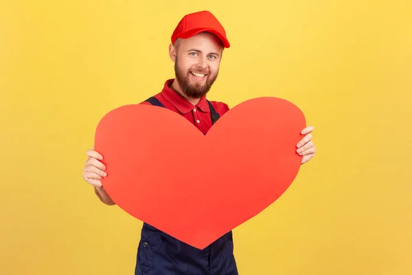 Portrait of pleased delighted worker man wearing blue overalls and red cap holding big red heart, fast gifts delivery during holidays. Indoor studio shot isolated on yellow background.