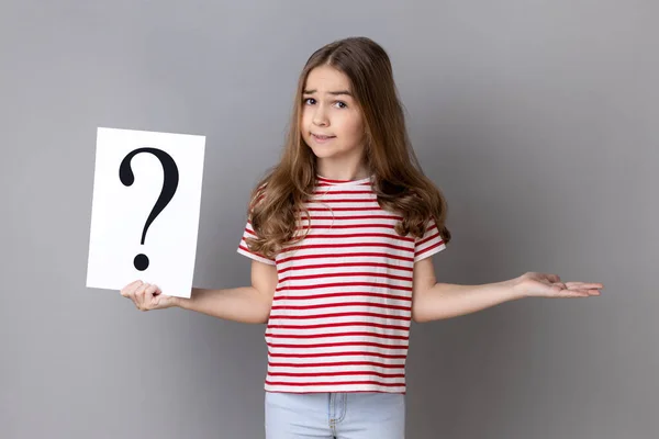 Portrait of little girl wearing striped T-shirt holding paper with question mark, making decision, not sure correct answer. Indoor studio shot isolated on gray background.