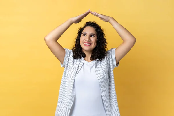 I\'m in safety. Portrait of woman with dark wavy hair raising hands showing roof gesture and smiling contentedly, dreaming of house, looking away. Indoor studio shot isolated on yellow background.