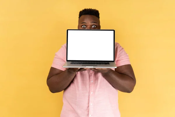 白い画面のノートパソコンで顔の半分を覆うピンクのシャツを着た男が 興奮した表情と大きな目でカメラを見ている 黄色の背景に隔離された屋内スタジオショット — ストック写真