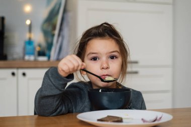 Güzel bir kız çocuğu siyah kaseden ekmek ve soğanla çorba içer. Mutfakta yemek yiyen bir çocuğun yaşam tarzı fotoğrafı..