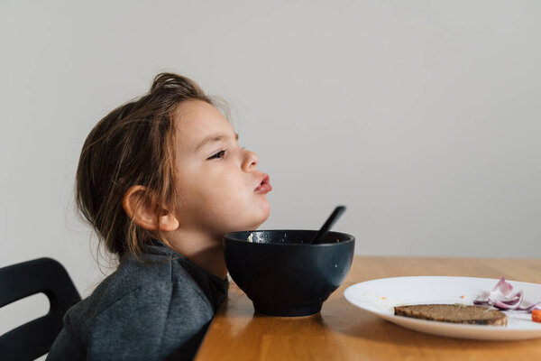 Unhappy child girl eats soup from black bowl with bread and onion. Lifestyle photo of kid in kitchen having a meal, Screaming kid. Picky eater