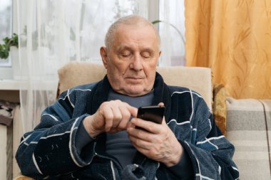 Akıllı telefonlu yaşlı bir adam koltukta oturuyor ve sosyal medyada kayıyor.. 