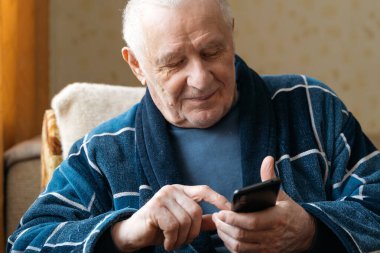 Akıllı telefonlu yaşlı bir adam koltukta oturuyor ve sosyal medyada kayıyor.. 