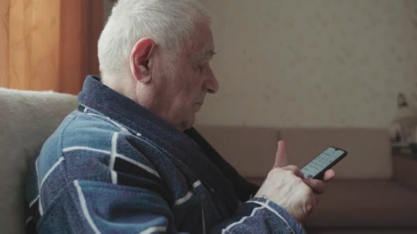 Homme âgé avec smartphone assis dans un fauteuil et faisant défiler les médias sociaux. — Video