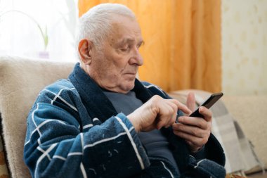 Akıllı telefonlu yaşlı bir adam koltukta oturuyor ve sosyal medyada kayıyor.. 