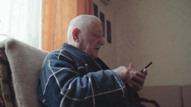 Homme âgé avec smartphone assis dans un fauteuil et faisant défiler les médias sociaux. — Video