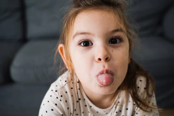 Oturma odasında kendini neşeli hissederken pijama giyen, dilini dışarı çıkaran küçük mutlu kızın portresi. — Stok fotoğraf