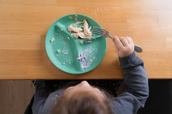 Menina criança sentada à mesa e colocando colher na boca enquanto come mingau atentamente e com apetite. Foto stock — Fotografia de Stock