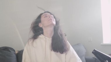 Genç beyaz kadın, saç kurutma makinesiyle evde, oturma odasındaki kanepede saç kurutuyor. 