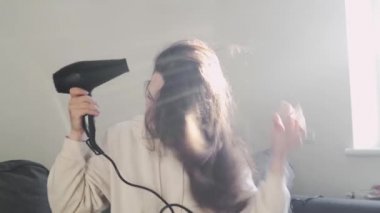 Genç beyaz kadın, saç kurutma makinesiyle evde, oturma odasındaki kanepede saç kurutuyor. 