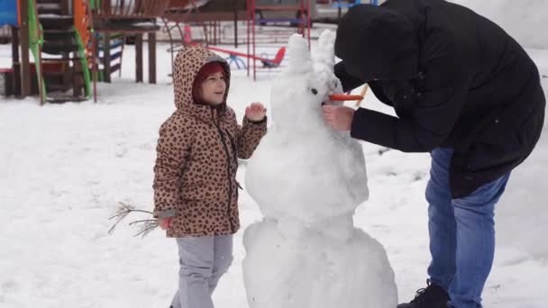 Far och barn leker på vintern snöig dag. Skapa snögubbe med familj på den urbana trädgården. — Stockvideo