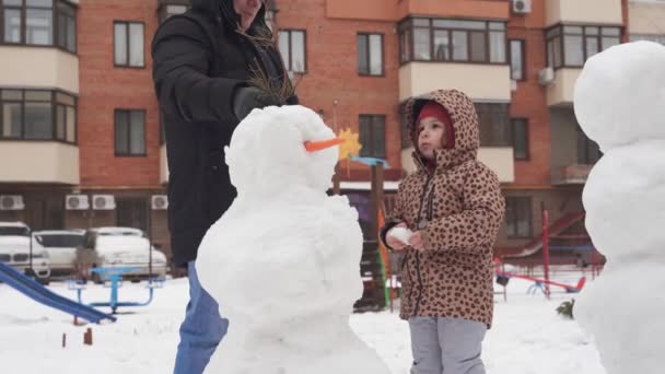 Far och barn leker på vintern snöig dag. Skapa snögubbe med familj på den urbana trädgården. — Stockvideo