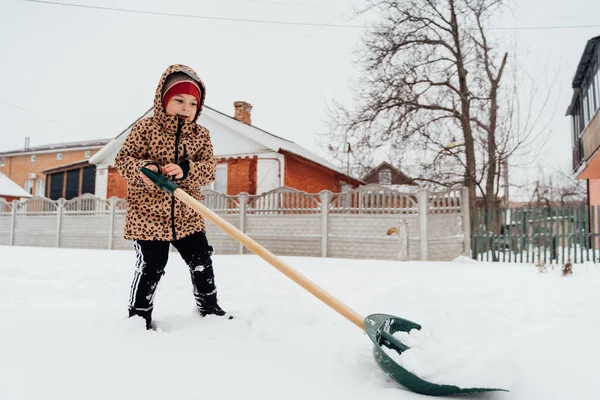 Vista de ángulo bajo de la niña ayudando a limpiar el camino de la nieve con la pala. Niño jugando en el jardín de invierno nevado — Foto de Stock