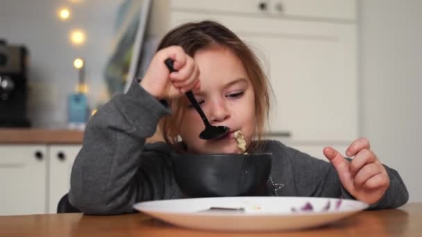 Mooi meisje eet soep uit zwarte kom met brood en ui. Lifestyle foto van kind in de keuken met een maaltijd. — Stockvideo