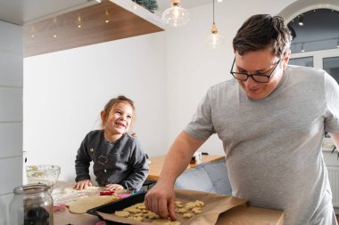 Tanımlanamayan bir adam ve kızının işleme tahtasında bisküvi kesicilerle şeker yaparken görüntülenmiş. Babam yemek tepsisine kurabiye koyuyor.